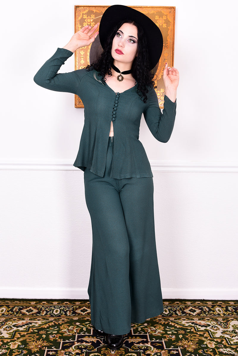 Woman in a vintage 1970s green trouser suit and black hat standing in a room with white wall.