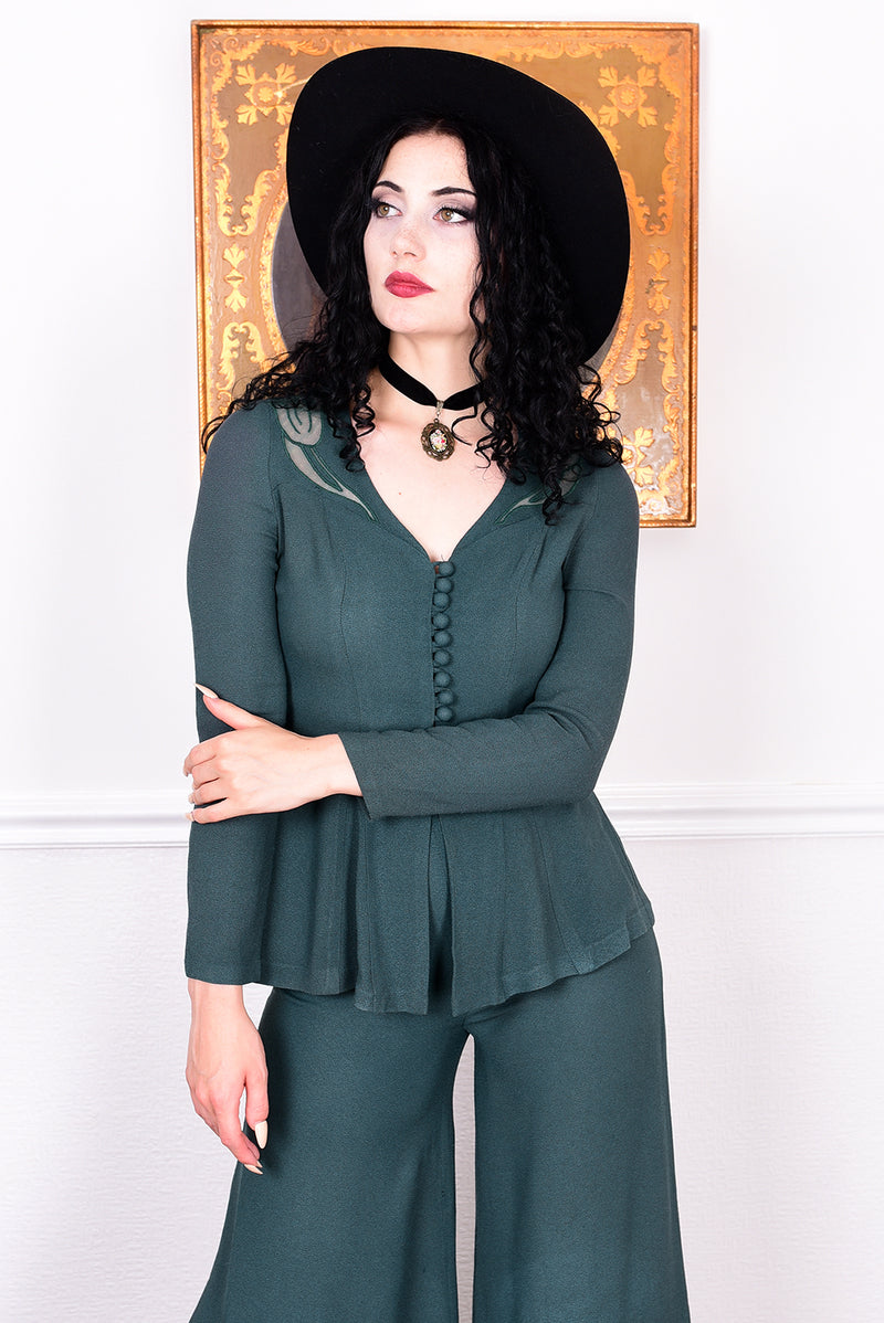 Woman in a vintage 1970s green trouser suit and black hat standing in a room with white wall.