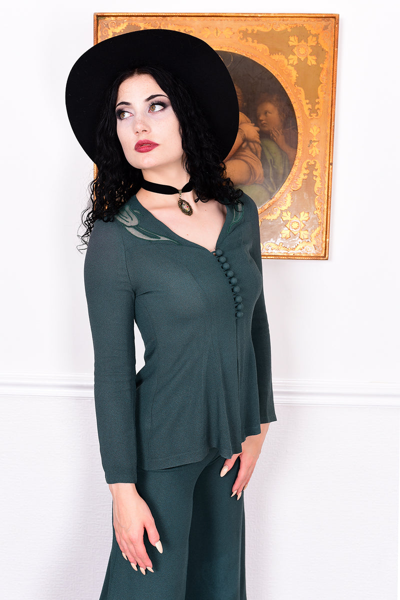 Woman in a vintage 1970s green trouser suit and black hat standing in a room with white wall.