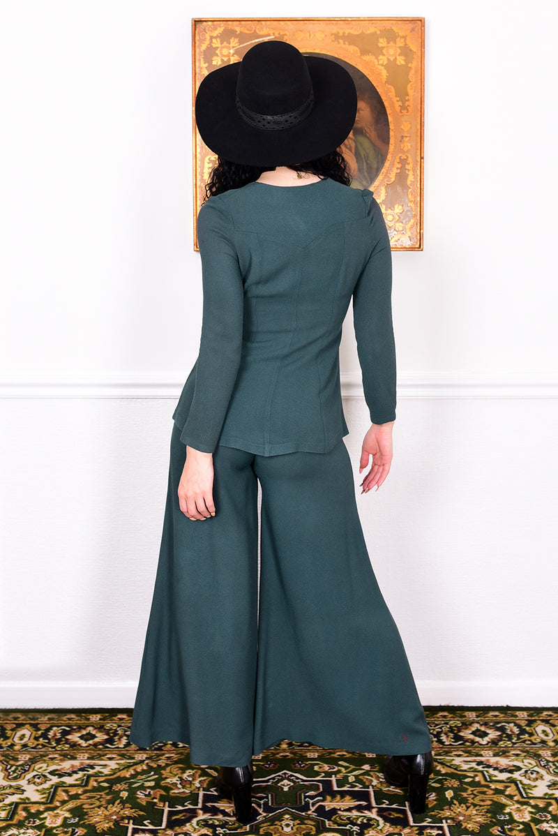 Woman in a vintage 1970s green trouser suit and black hat standing in a room with white wall.