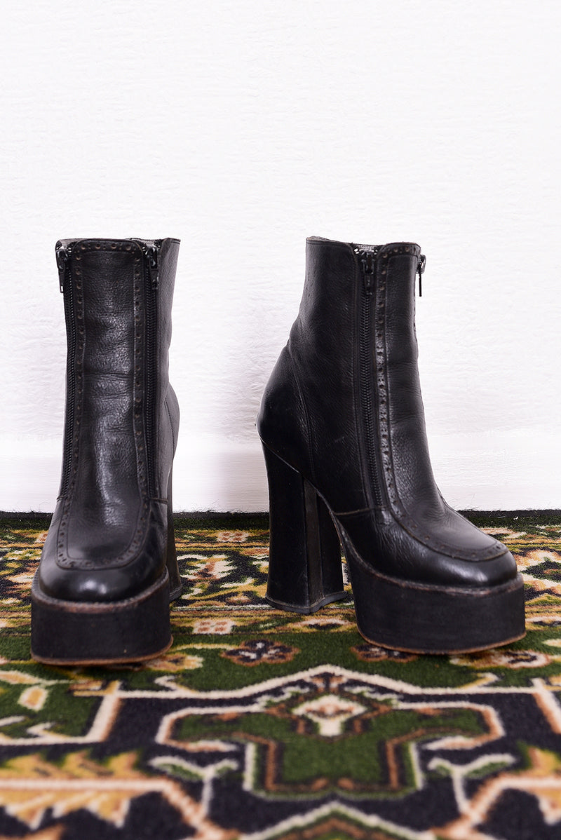 Pair of black vintage 1990s platform boots on a patterned rug with a white wall background.