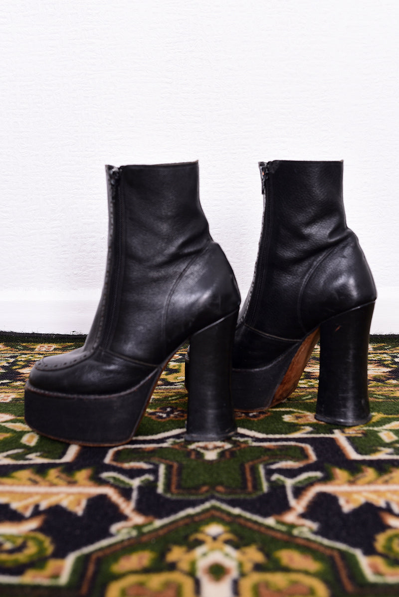 Pair of black vintage 1990s platform boots on a patterned rug with a white wall background.