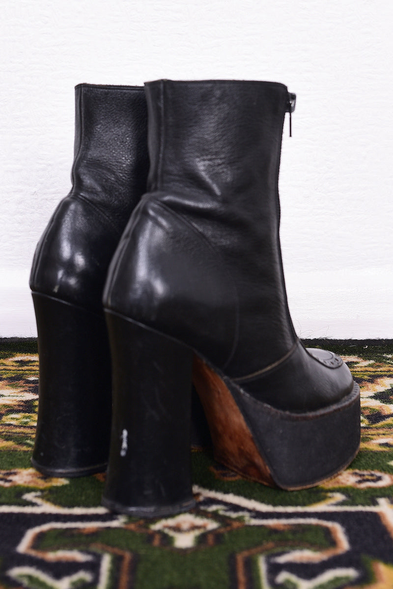Pair of black vintage 1990s platform boots on a patterned rug with a white wall background.