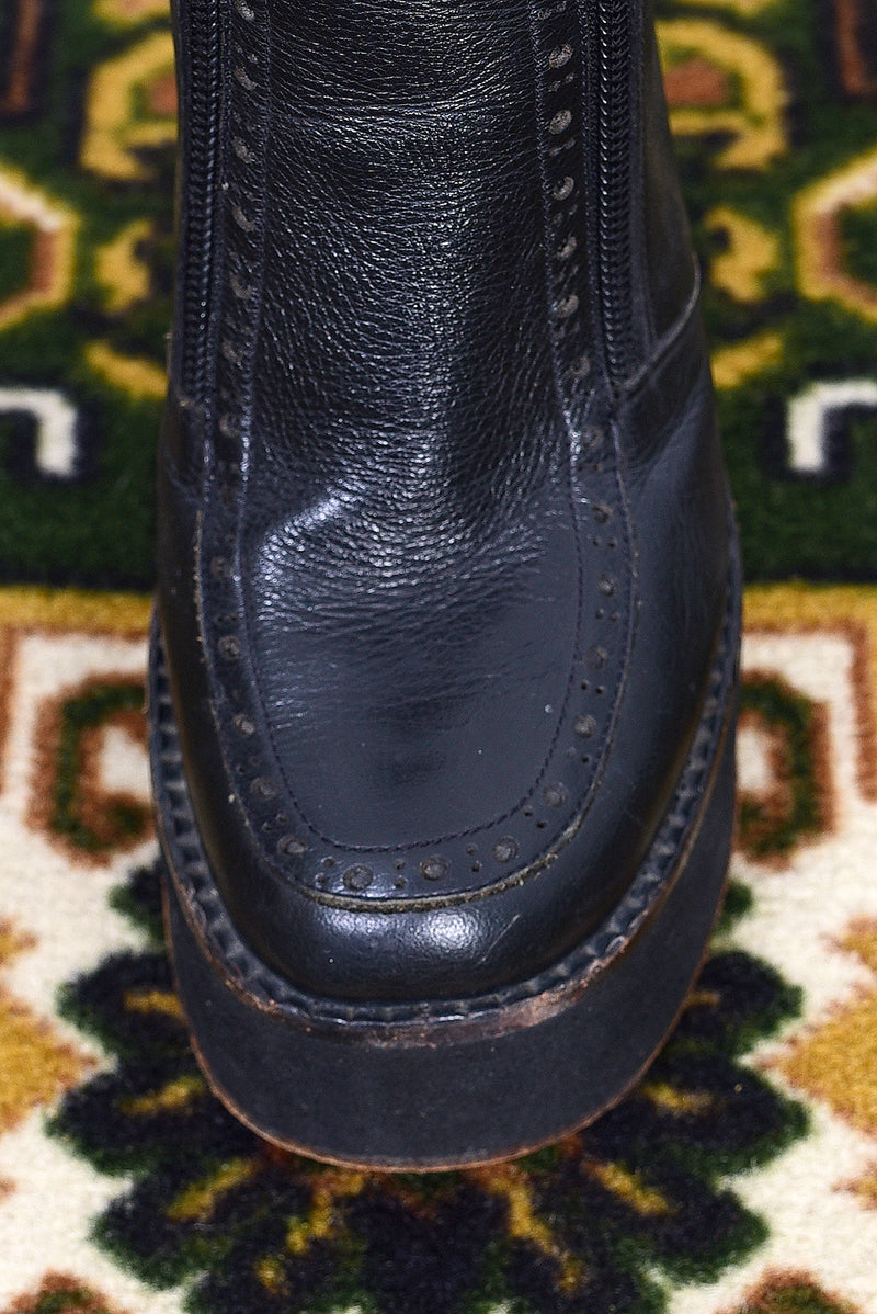 Pair of black vintage 1990s platform boots on a patterned rug with a white wall background.