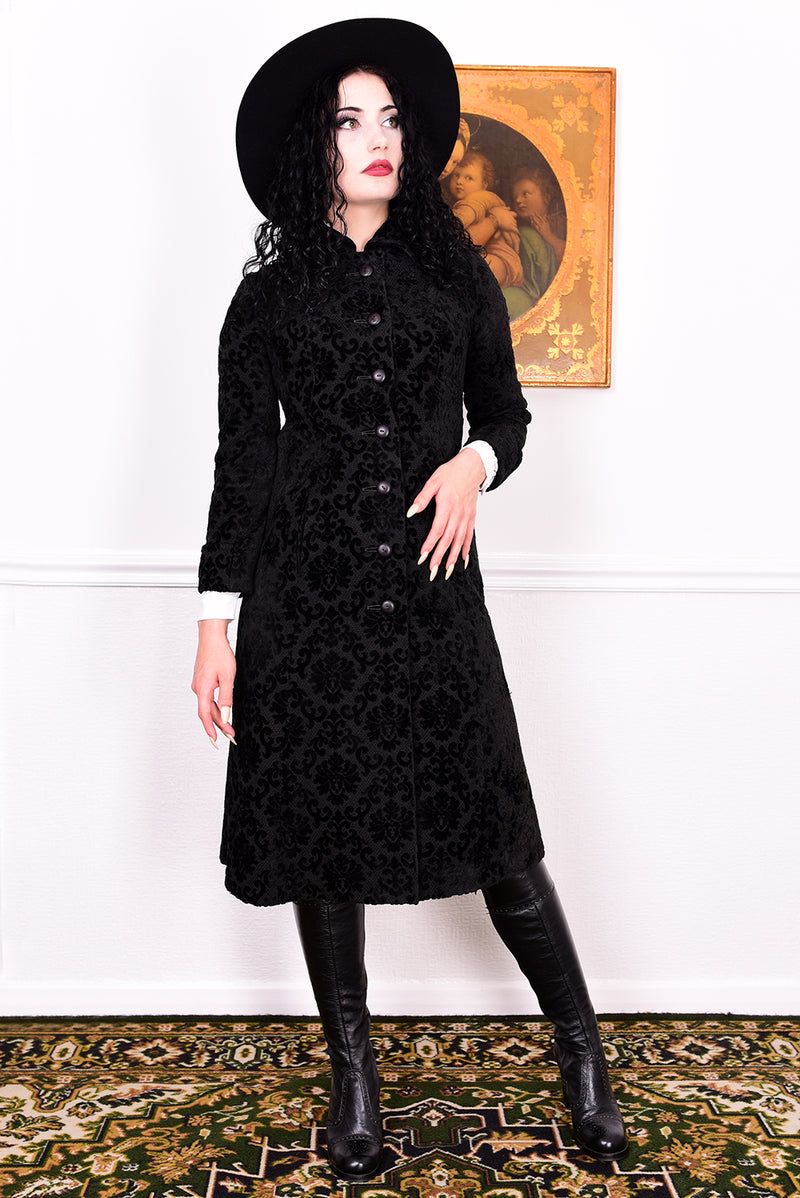 Woman in a vintage 1960s black damask coat and hat standing against a white wall.