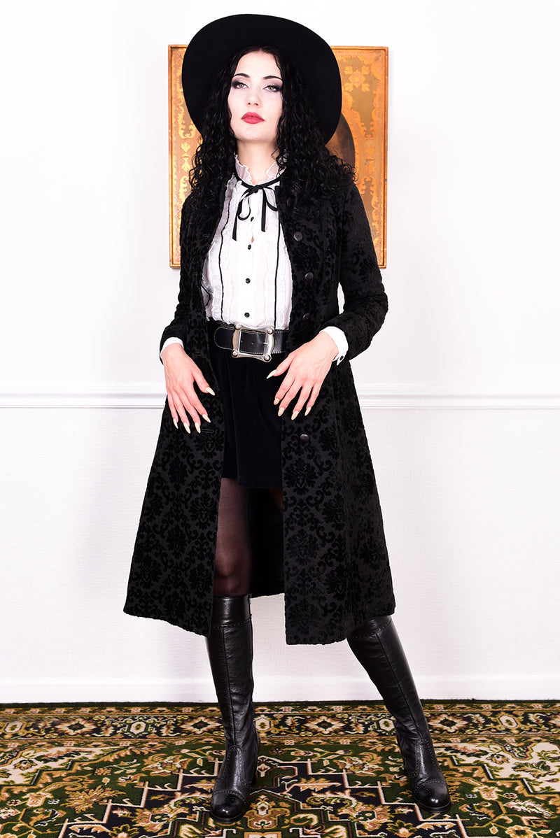 Woman in a vintage 1960s black damask coat and hat standing against a white wall.