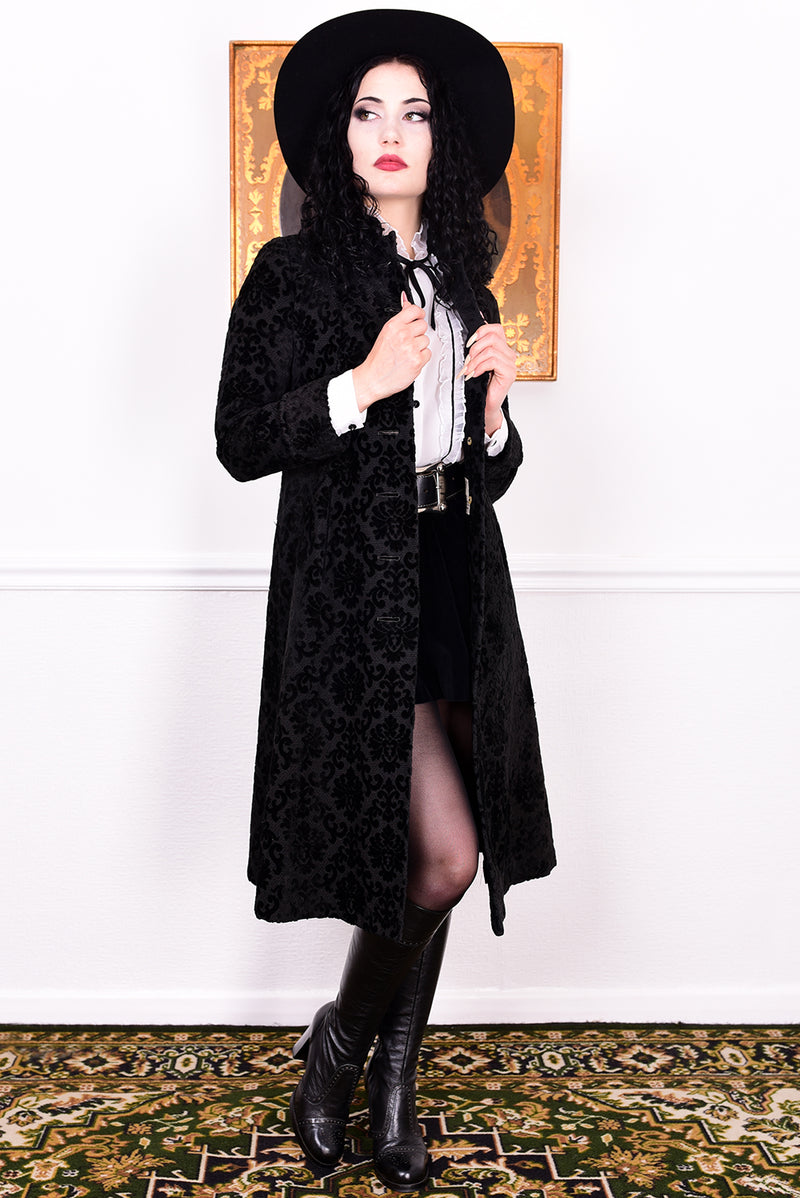 Woman in a vintage 1960s black damask coat and hat standing against a white wall.