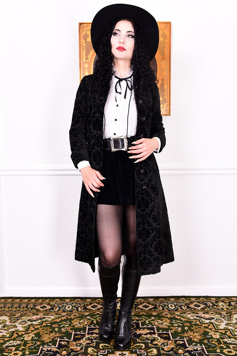 Woman in a vintage 1960s black damask coat and hat standing against a white wall.