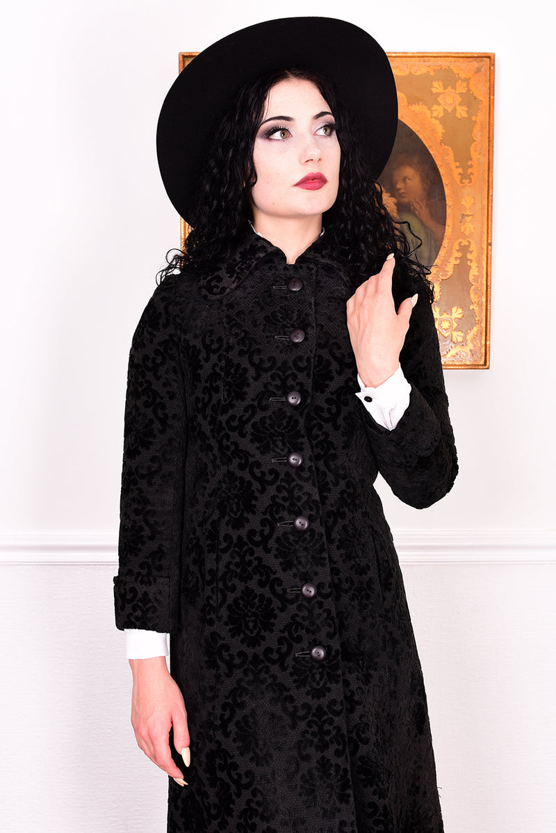 Woman in a vintage 1960s black damask coat and hat standing against a white wall.