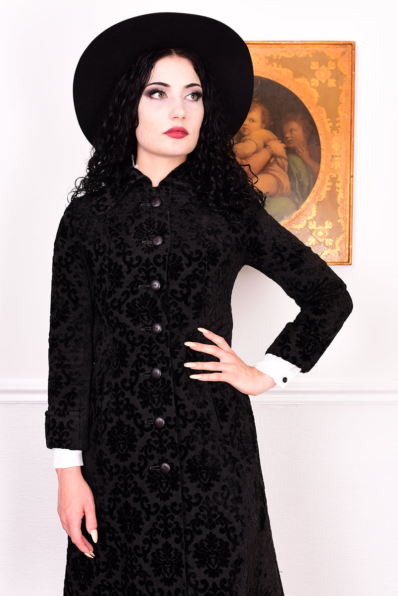 Woman in a vintage 1960s black damask coat and hat standing against a white wall.
