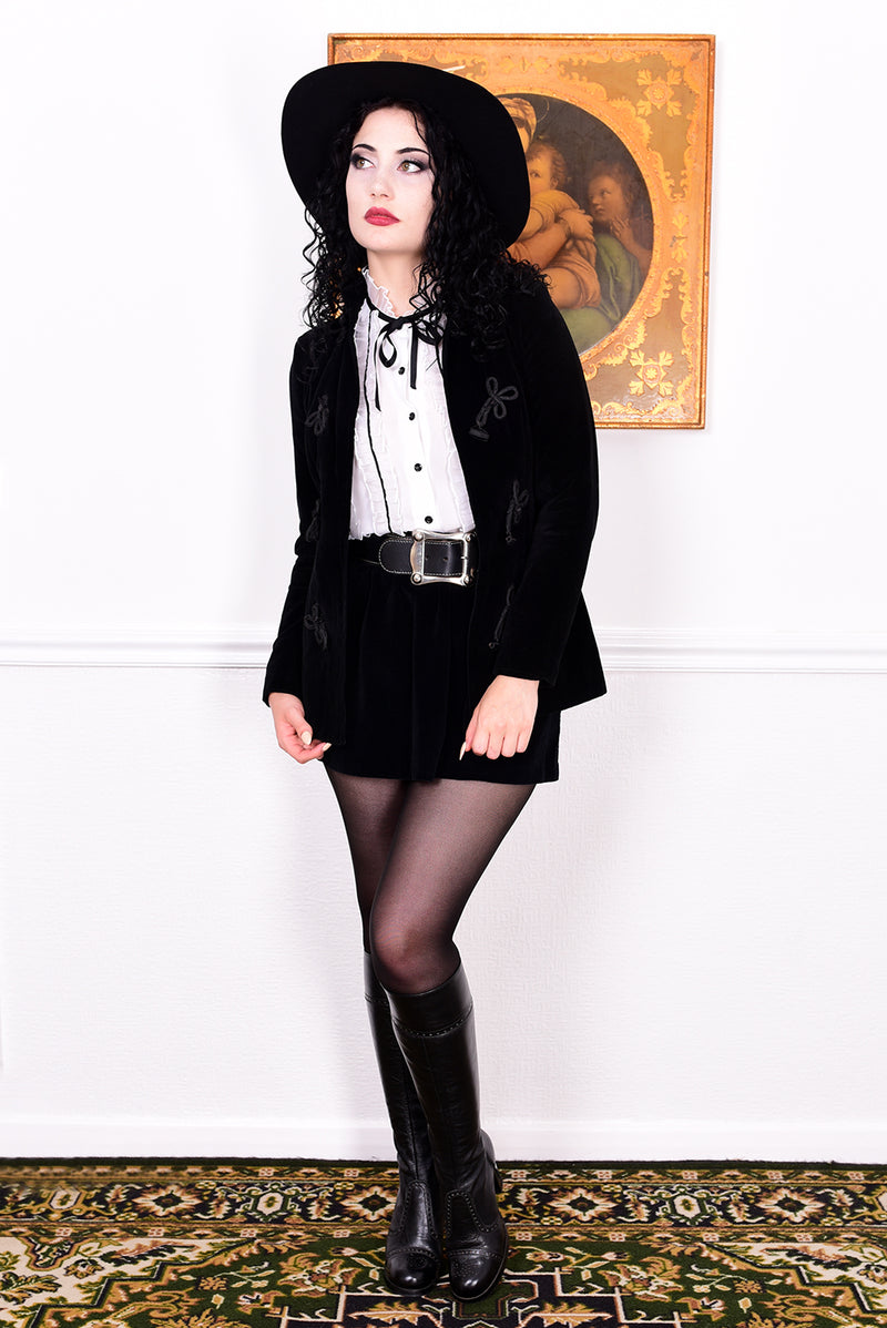 Woman wearing a vintage 1970s black velvet jacket with frog closures in front of a white wall.