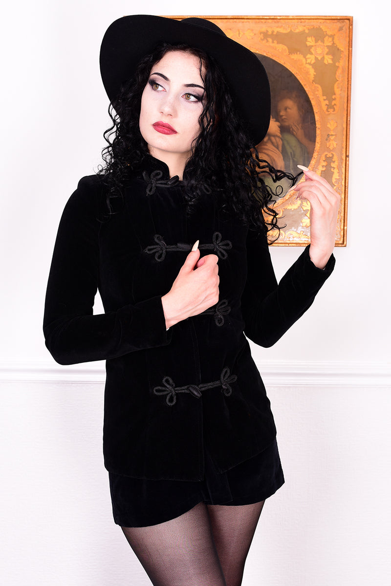 Woman wearing a vintage 1970s black velvet jacket with frog closures in front of a white wall.