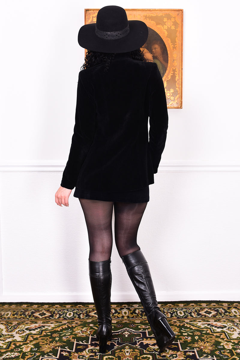 Woman wearing a vintage 1970s black velvet jacket with frog closures in front of a white wall.