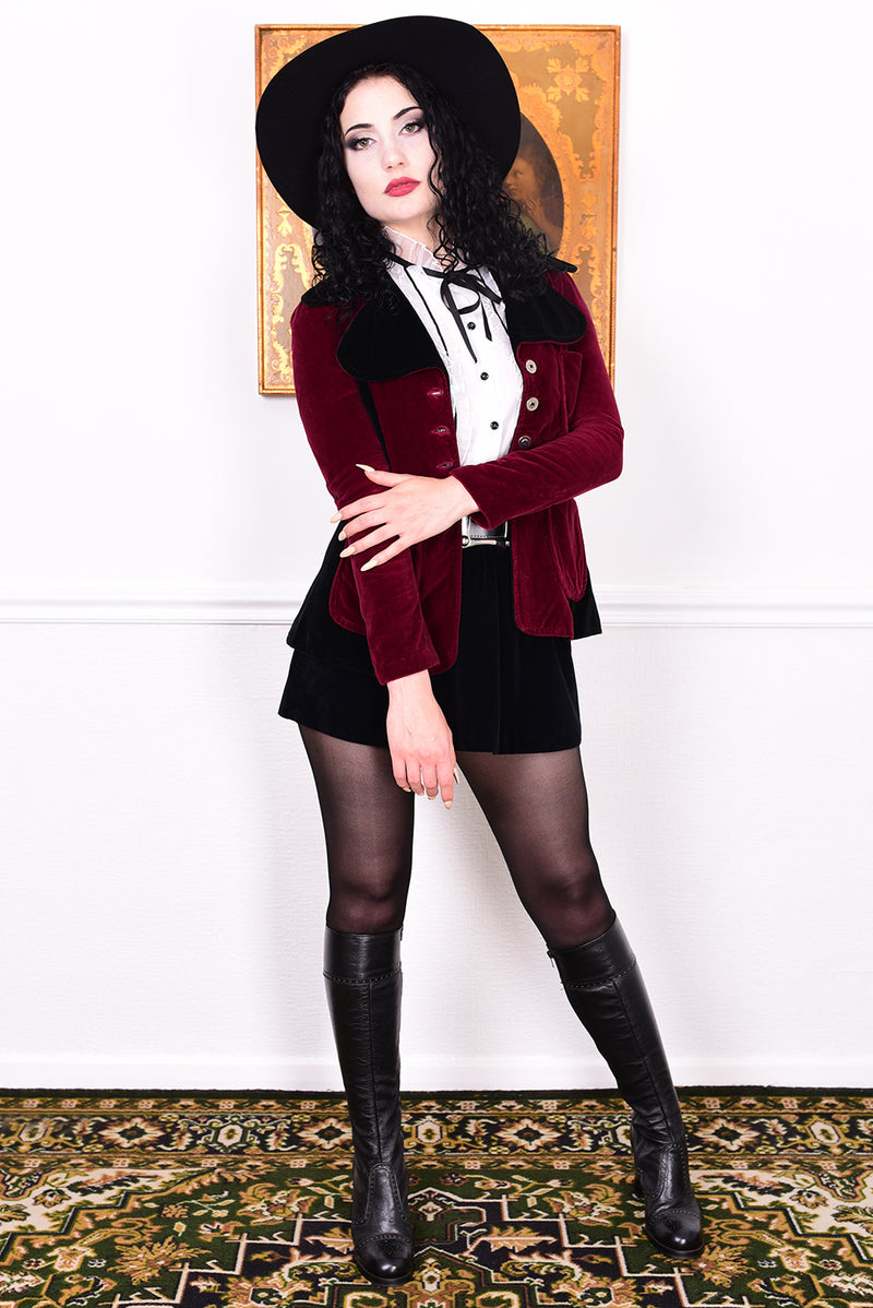 Woman wearing a vintage 1960s black and burgundy velvet jacket with a black hat, standing in front of a white wall.