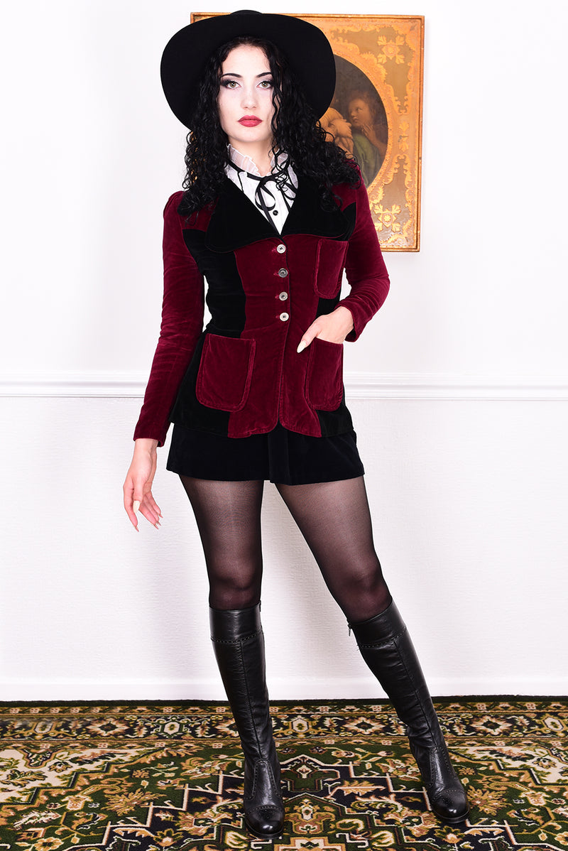 Woman wearing a vintage 1960s black and burgundy velvet jacket with a black hat, standing in front of a white wall.