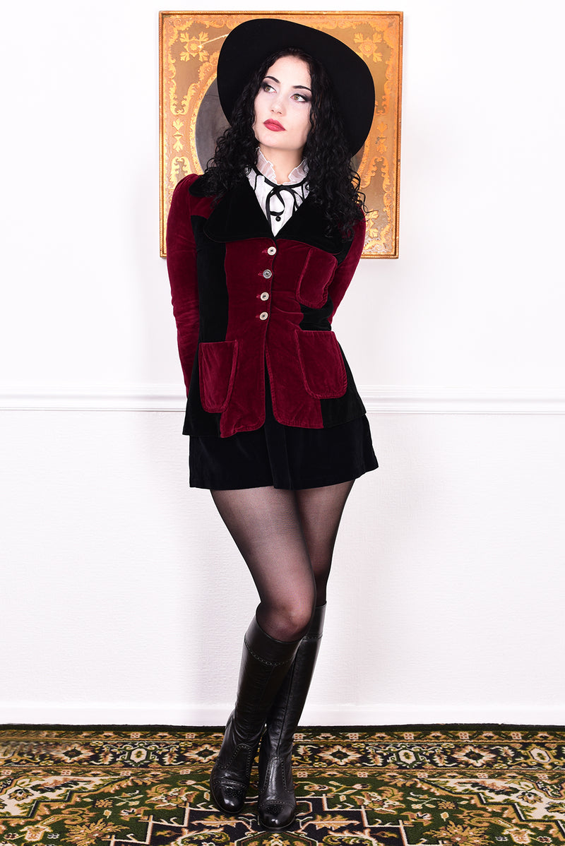 Woman wearing a vintage 1960s black and burgundy velvet jacket with a black hat, standing in front of a white wall.