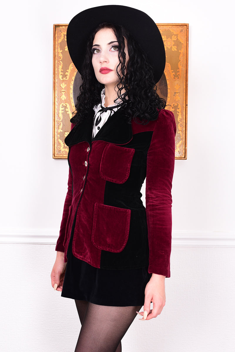 Woman wearing a vintage 1960s black and burgundy velvet jacket with a black hat, standing in front of a white wall.