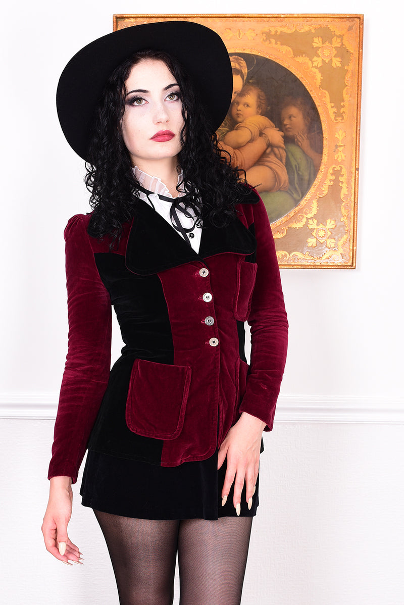 Woman wearing a vintage 1960s black and burgundy velvet jacket with a black hat, standing in front of a white wall.