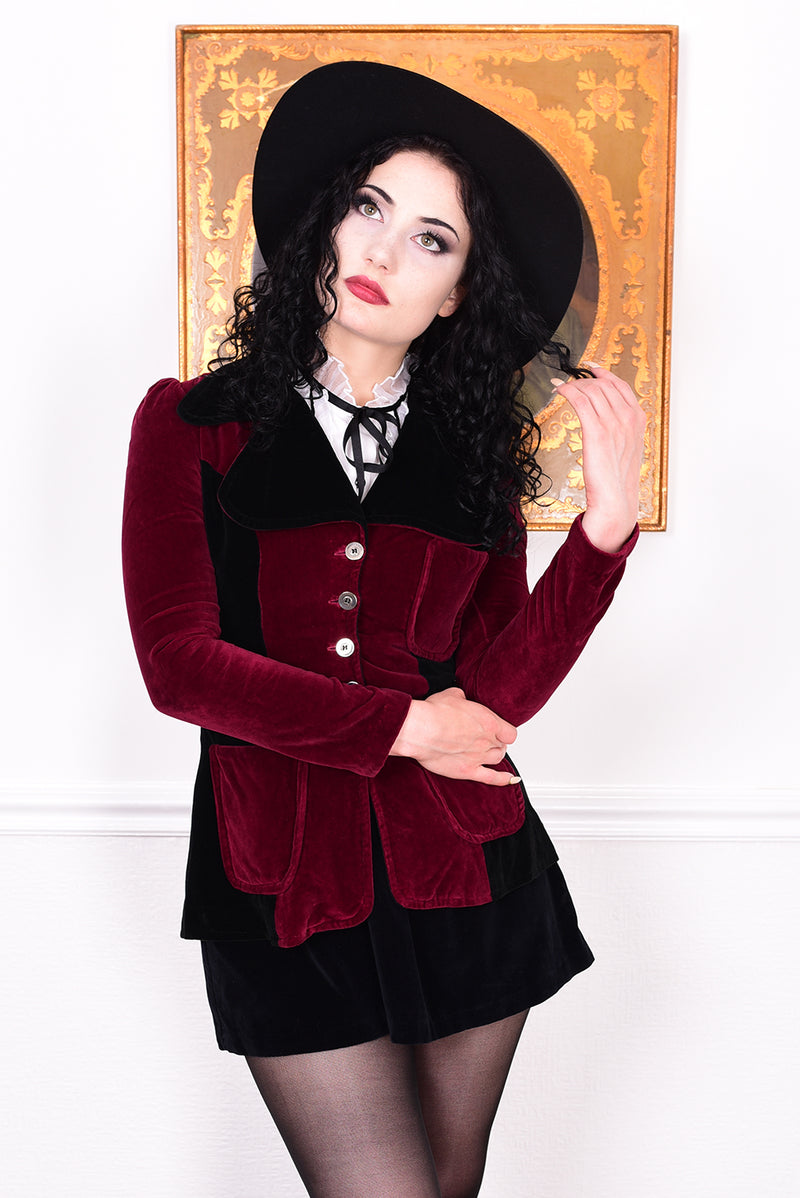 Woman wearing a vintage 1960s black and burgundy velvet jacket with a black hat, standing in front of a white wall.