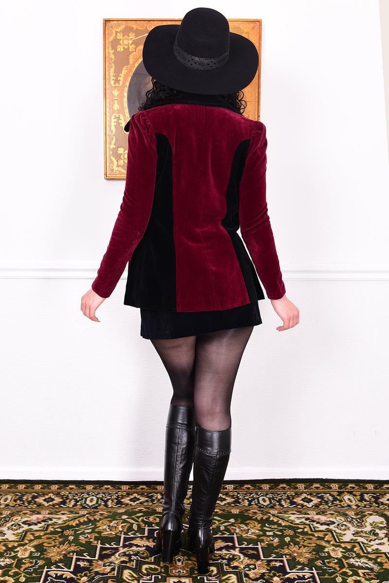 Woman wearing a vintage 1960s black and burgundy velvet jacket with a black hat, standing in front of a white wall.