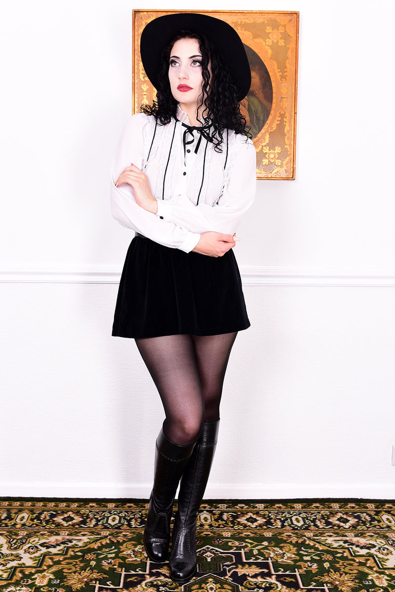 Woman in a white blouse, black skirt, and black boots standing in a room with a decorative rug and wall art.