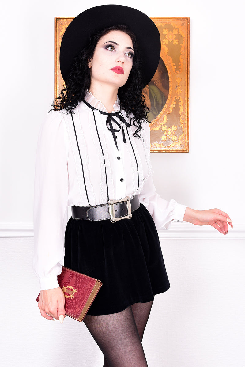 Woman in a white blouse with black trim, black skirt, and black hat holding a red book against a white wall.