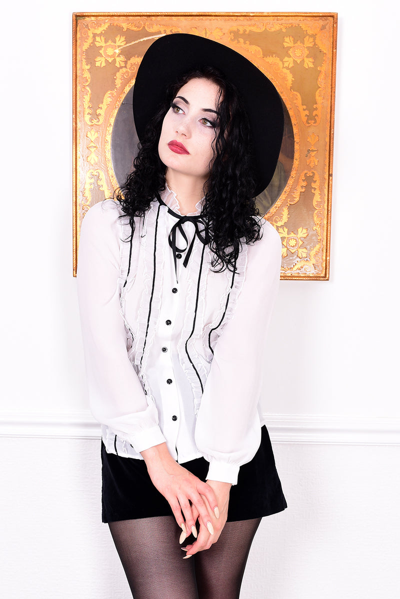 Woman wearing a white blouse with black buttons, black skirt, and black stockings in front of a decorative wall.