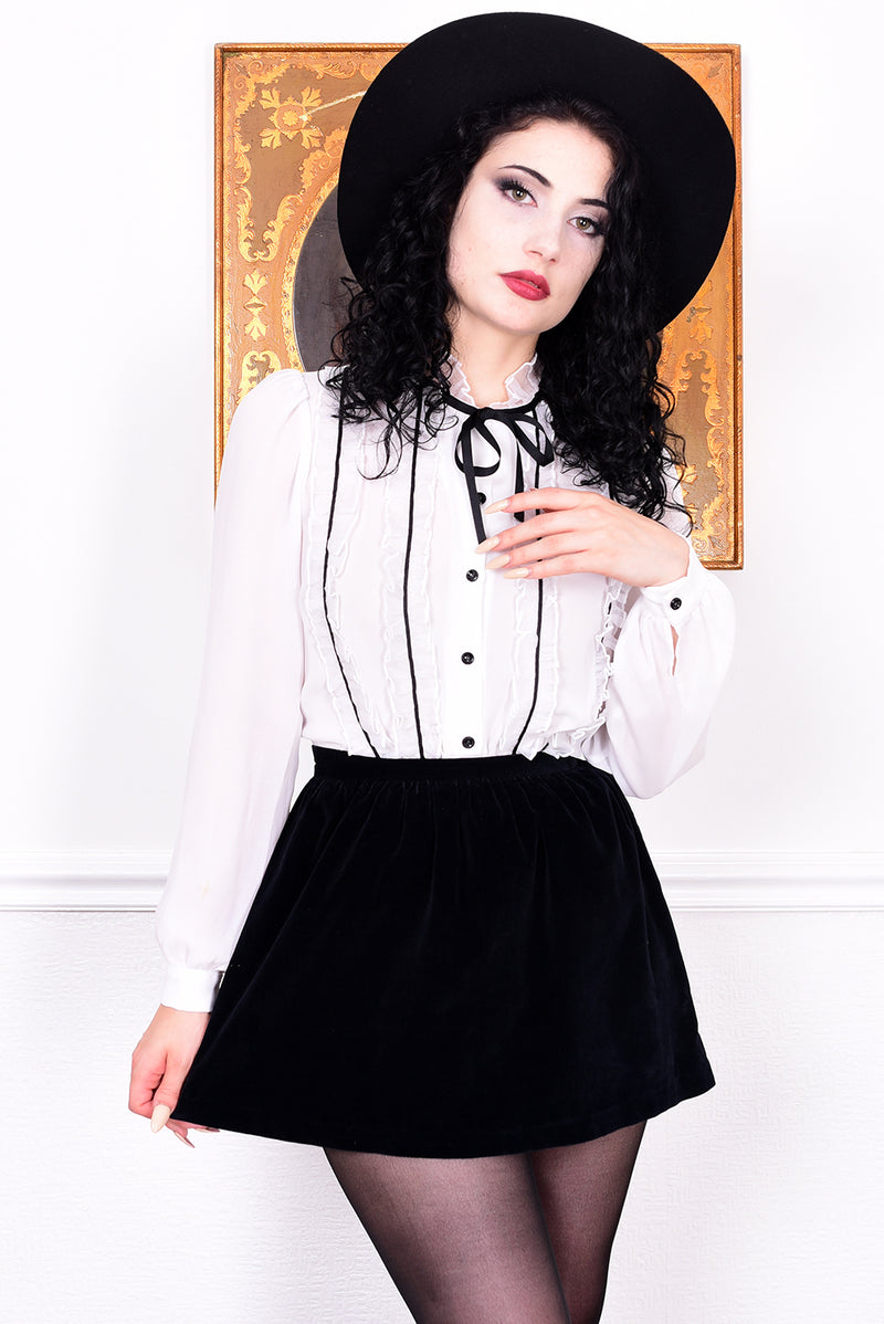 Woman wearing a white blouse with black buttons and a black skirt, standing against a white wall.