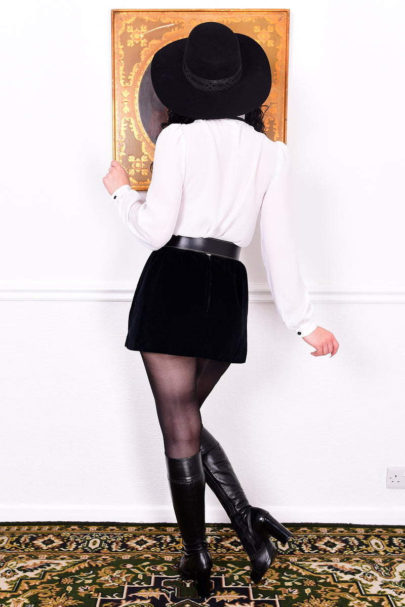 Woman wearing a black hat, white shirt, black skirt, and black boots in a room with a decorative rug.