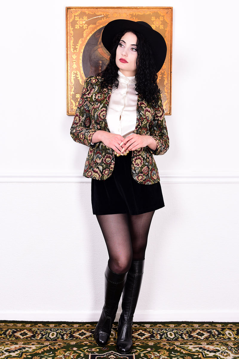 Woman in vintage 1970s tapestry floral blazer, black skirt, and boots standing in front of a white wall.