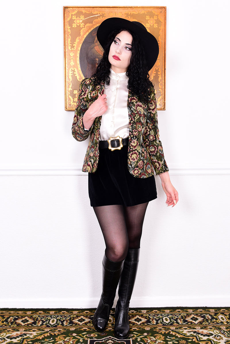 Woman in vintage 1970s tapestry floral blazer, black skirt, and boots standing in front of a white wall.