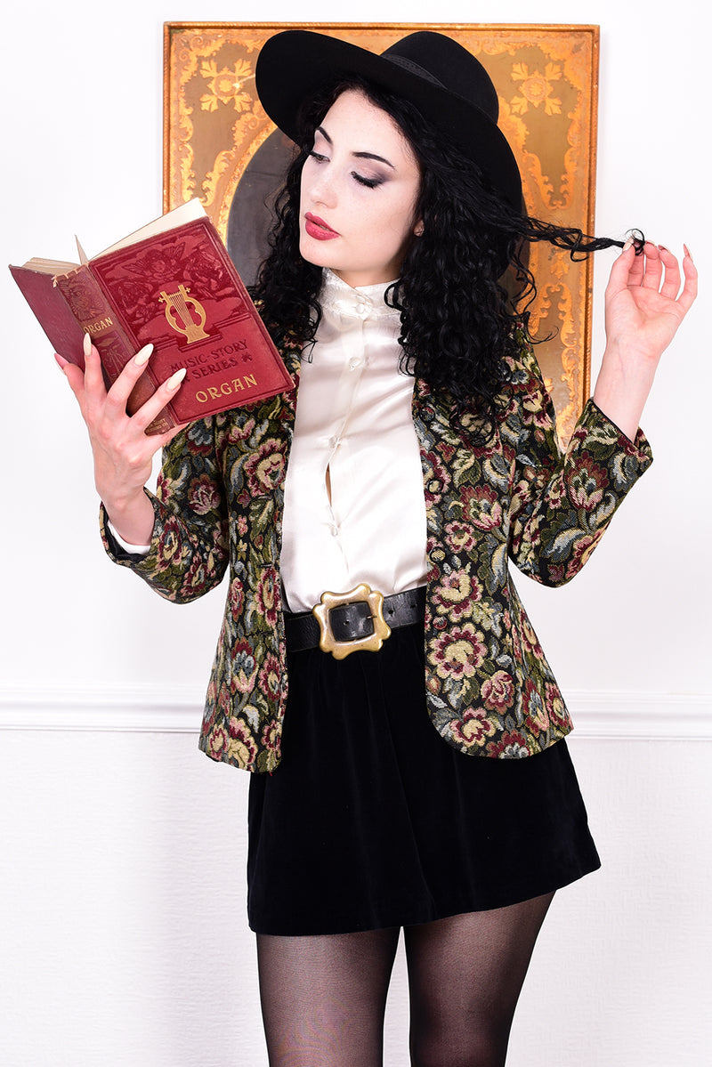 Woman in vintage 1970s tapestry floral blazer, black skirt, and boots standing in front of a white wall.