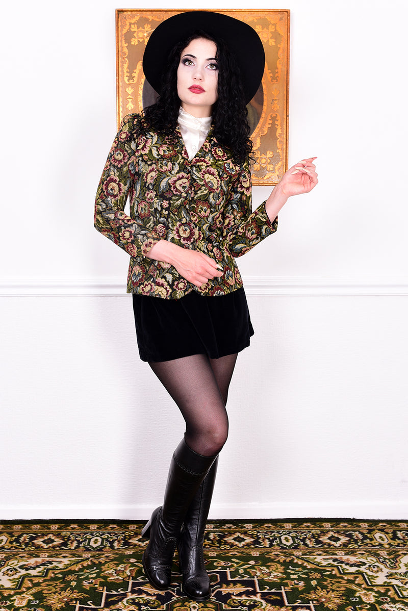 Woman in vintage 1970s tapestry floral blazer, black skirt, and boots standing in front of a white wall.
