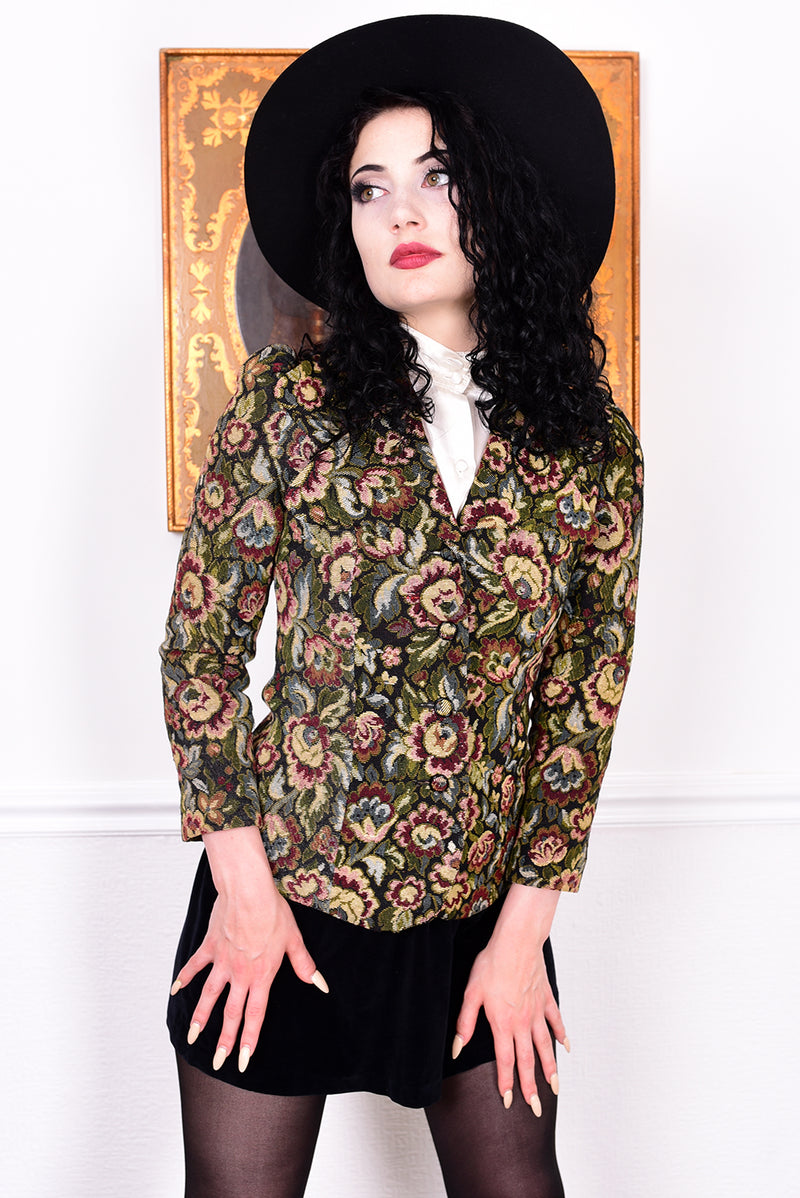 Woman in vintage 1970s tapestry floral blazer, black skirt, and boots standing in front of a white wall.