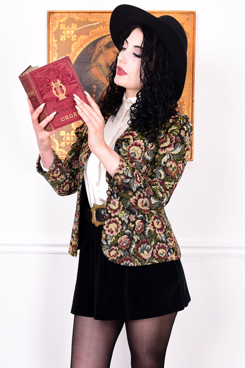 Woman in vintage 1970s tapestry floral blazer, black skirt, and boots standing in front of a white wall.