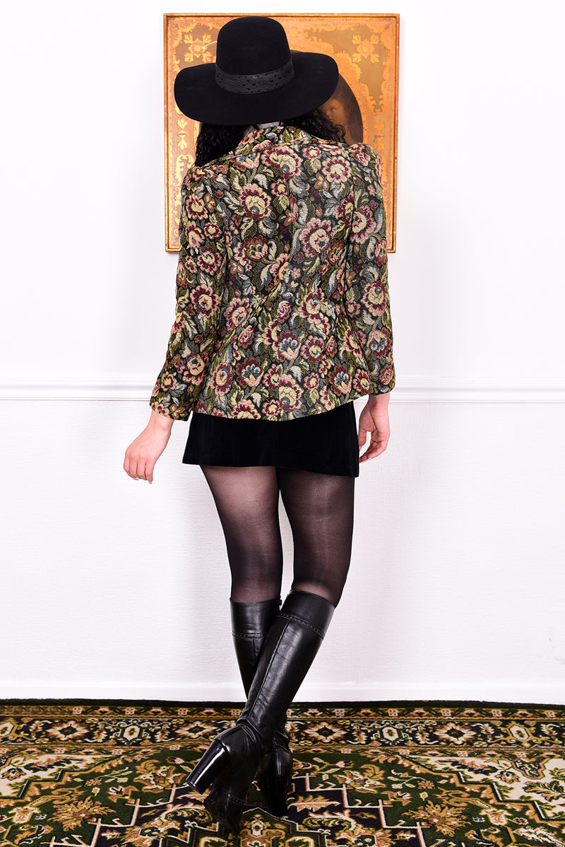 Woman in vintage 1970s tapestry floral blazer, black skirt, and boots standing in front of a white wall.